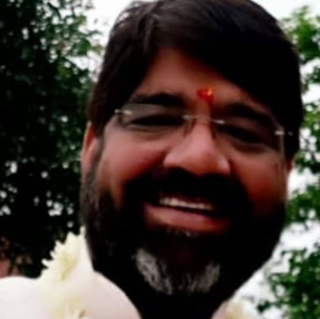 Smiling Indian spiritual guru wearing glasses and a tilak on his forehead, with a garland around his neck, standing outdoors with trees in the background.